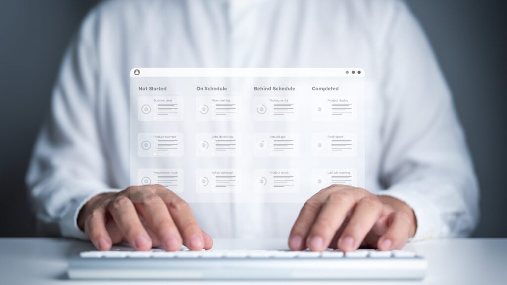 A man, who appears to be in his late 30s, with light skin and short brown hair, is shown from the chest down, wearing a white collared shirt. He is seated at a white desk with his hands positioned over a white keyboard. In front of him, a transparent, holographic interface displays a project management dashboard. The dashboard is divided into four columns: "Not Started," "On Schedule," "Behind Schedule," and "Completed." Each column contains several cards, each representing a task with a title and a progress bar. The interface has a clean, minimalist design with a light color palette and soft, diffused lighting. The background is a dark, out-of-focus gray. The image is a digital illustration with a shallow depth of field, focusing on the man's hands and the holographic interface.